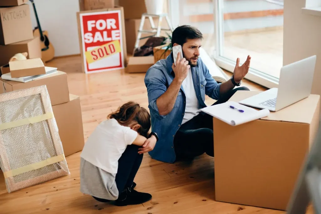 Man on phone looking frustrated while sitting on cardboard boxes, with a child sitting nearby, in a room filled with moving boxes, reflecting tenant challenges and legal issues in Bouldin Creek.