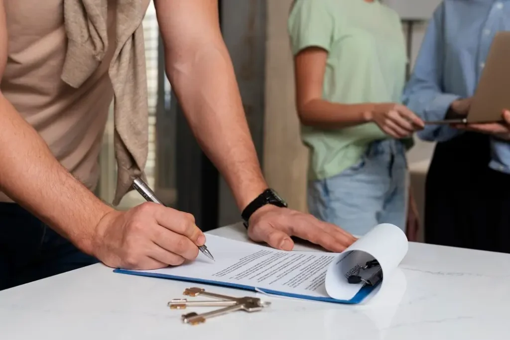 Person signing a lease agreement with keys on the table, while two others discuss terms, illustrating tenant law services in Bryker Woods.