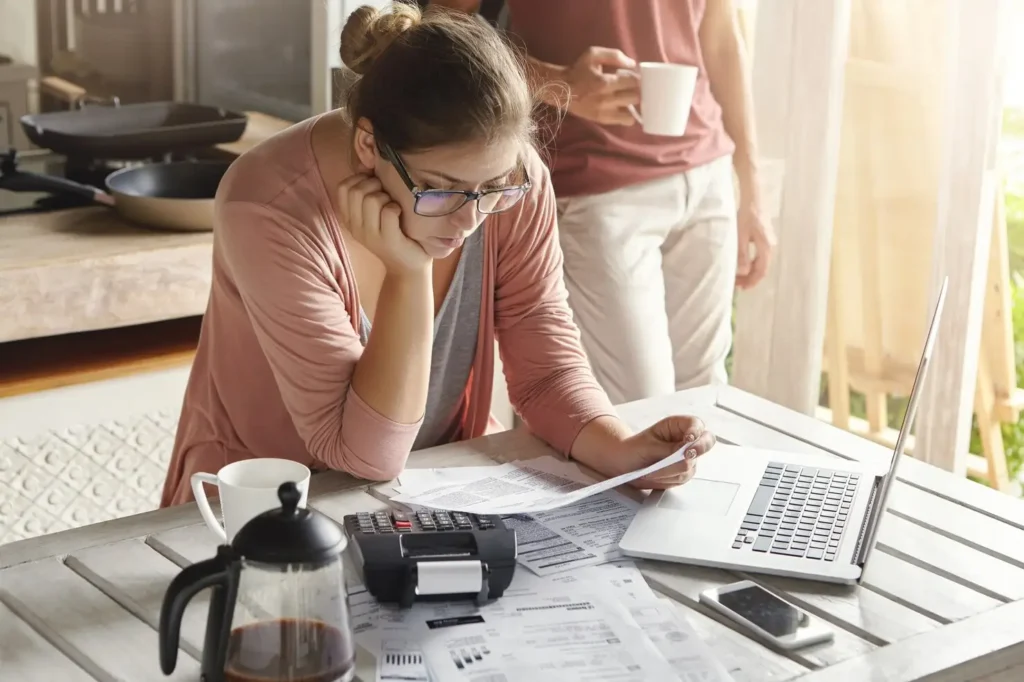 Woman reviewing documents and bills at a table with a laptop and coffee pot, while a man stands nearby holding a cup, illustrating tenant rights and financial concerns in Central Austin.