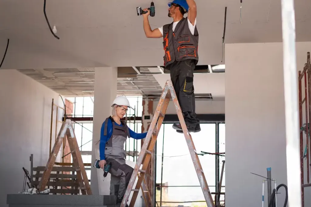 Jester Estates Two construction workers, one on a ladder using a drill and the other on a lower ladder holding a tool, working on ceiling repairs in an interior space, illustrating tenant rights related to repair and maintenance enforcement in Jester Estates.