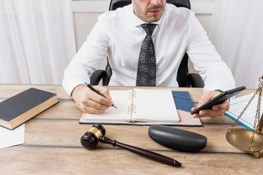 Lawyer in white shirt and tie writing in notebook, holding smartphone, with gavel, law books, and scales of justice on desk, representing legal services for tenants and landlords in Johnson County.