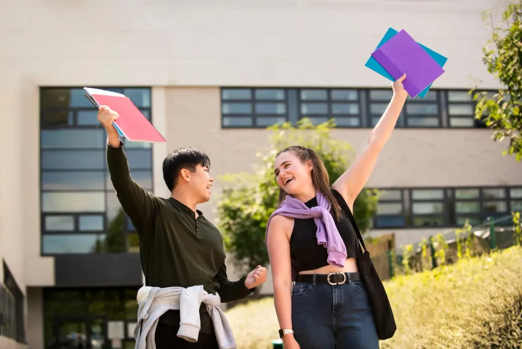 Two happy students celebrating outdoors, holding colorful folders, in front of a modern building, symbolizing educational success and tenant rights advocacy in Old West Austin.