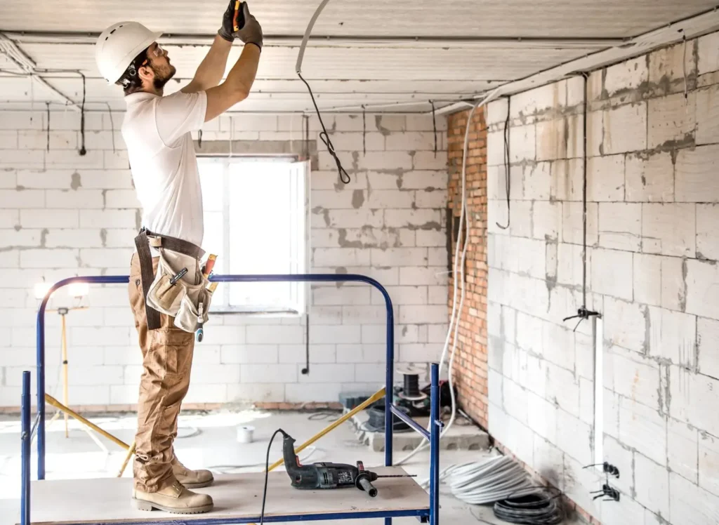 Construction worker on ladder installing ceiling fixtures in a partially renovated room with exposed walls and electrical wiring.